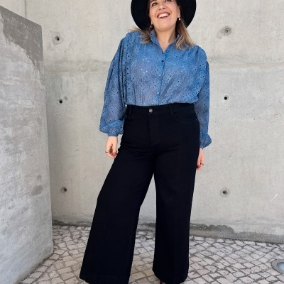 Mulher com blusa azul, calças pretas largas e chapéu preto em frente a parede de cimento.