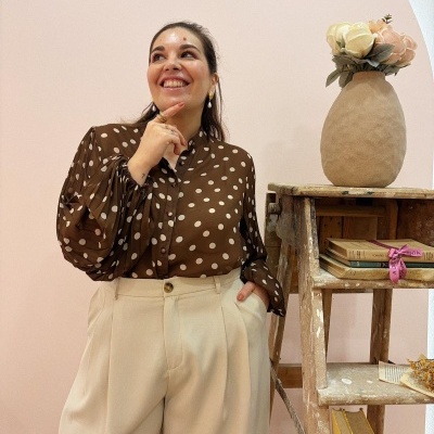 Mulher com camisa castanha de bolinhas brancas e calças bege junto a escadote com livros e vaso de flores