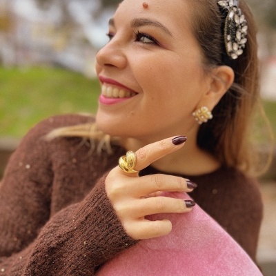 Mulher sorridente com anel dourado, brincos perolados, tiara com pedras e suéter castanho segurando almofada rosa veludo