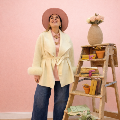 Mulher com casaco creme e chapéu rosa junto a escada com livros e flores.