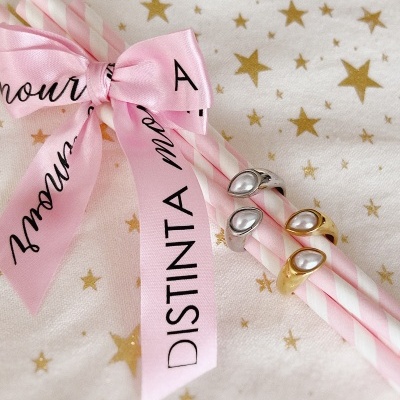 Anéis com pedras em forma de gota em canudos rosa e branco, laço rosa com texto 'DISTINTA', fundo branco com estrelas douradas.