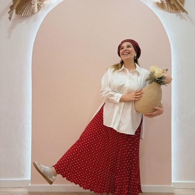 Mulher com camisa branca, saia vermelha de bolinhas, boina vermelha e sapatilhas, segurando vaso com flores num arco decorado.