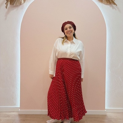 Mulher vestida com boina vermelha, camisa branca e saia longa vermelha de bolinhas brancas, em arco cor-de-rosa com decoração floral seca.