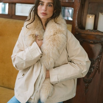 Mulher com casaco bege com gola de pelo bege claro, sentada em sofá amarelo em ambiente interior.