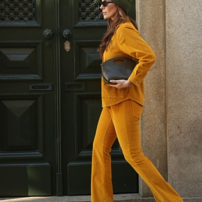 Mulher de perfil com conjunto amarelo, botas pretas, chapéu verde com pompom e mala castanha junto a porta verde.