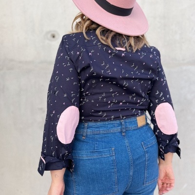 Mulher de costas com blusa preta com padrões e cotoveleiras rosas, calças de ganga azuis e chapéu rosa