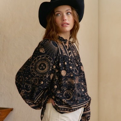 Blusa preta com estampados celestes em dourado, calças brancas e chapéu preto em ambiente interior.