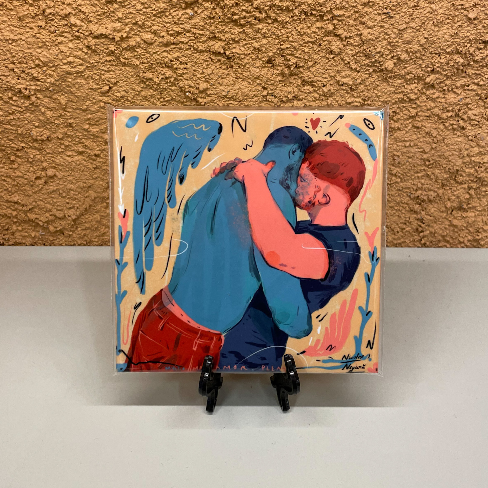 AZULEJO "ABRAÇO" NICOLAE NEGURA Ilustração de dois homens a abraçar-se, um azul com asas e outro ruivo, moldura em suporte preto.