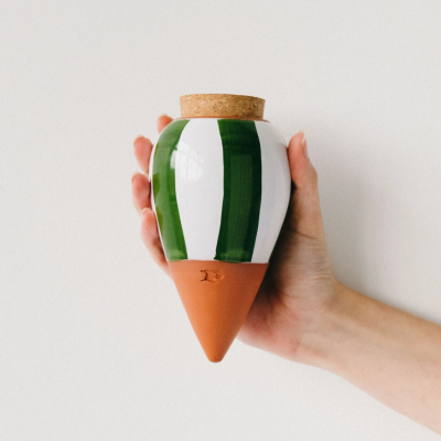 Frasco cerâmico branco com riscas verdes e base em terracota, tampa de cortiça, segurado por mão