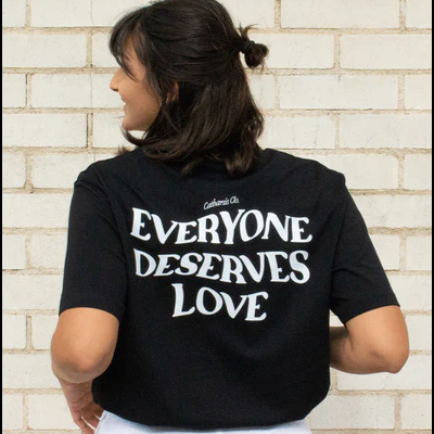 Pessoa de costas usando uma t-shirt preta com texto branco e calças brancas, diante de uma parede de tijolos clara