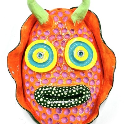 Máscara cerâmica decorativa laranja, olhos amarelos/azuis, boca verde com pontos brancos, chifres verdes