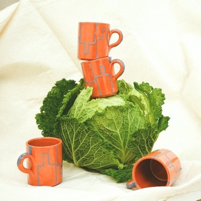 Quatro chávenas de cerâmica cor de laranja com padrão azul empilhadas e distribuídas sobre uma couve e tecido bege claro