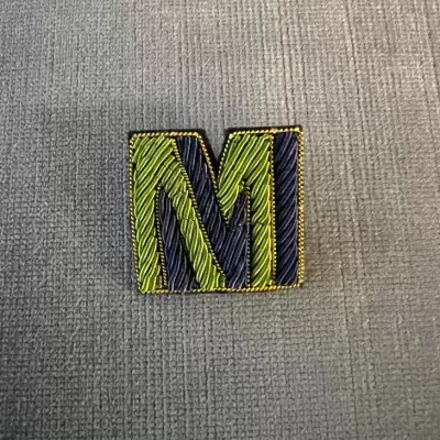 Patch bordado da letra M verde e preto com contorno amarelo sobre tecido cinzento