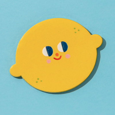 Prato cerâmico amarelo em forma de limão com rosto sorridente sobre fundo azul