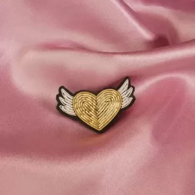 Broche de coração dourado com asas brancas sobre tecido rosa acetinado
