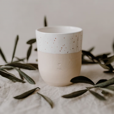 Copo cerâmico bege e branco com texto '❤️ HIS' sobre tecido com ramos verdes