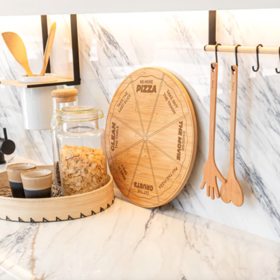 Bancada de cozinha com tábua de madeira redonda gravada, potes de vidro com cereais, copos cerâmicos e utensílios de madeira pendurados