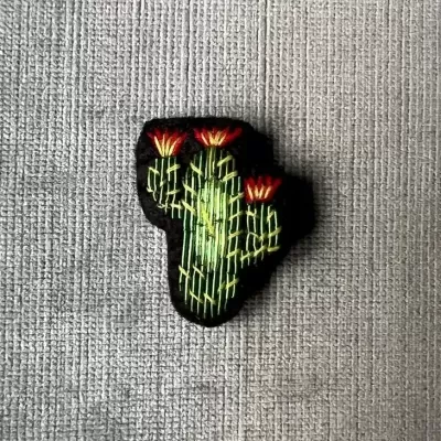 Broche tecido em forma de cacto com flores vermelhas sobre tecido cinzento