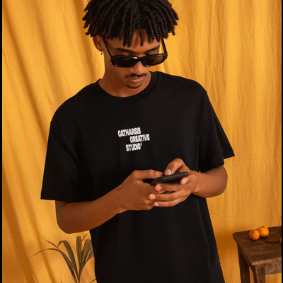 Homem com t-shirt preta 'CATHARSIS CREATIVE STUDIÓ' e óculos de sol em fundo amarelo com planta e mesa com tangerinas