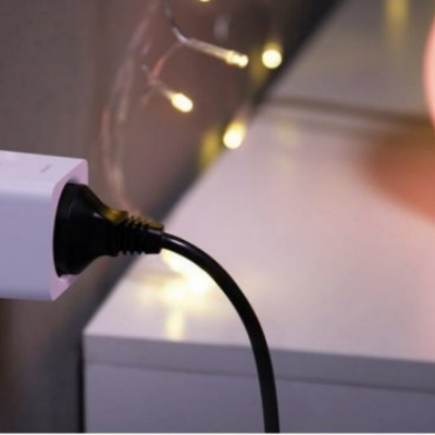 Tomada elétrica com adaptador branco e cabo preto junto a mesa branca com lâmpada e luzes decorativas