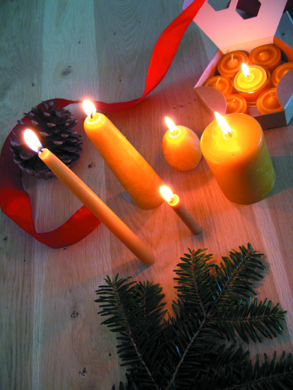 Cinco velas acesas com fita vermelha, pinha e ramo verde sobre madeira clara.