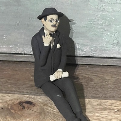 Figura decorativa masculina vestida de preto sentada num chão de madeira com fundo cinza