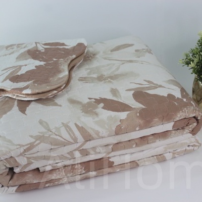 Colcha de padrão floral bege e castanho dobrada ao lado de almofada e vaso dourado com planta