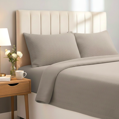 Conjunto de cama cinzento claro com almofadas e edredão, cabeceira branca acolchoada, criado-mudo com jarra de flores, livro, chávena e candeeiro.