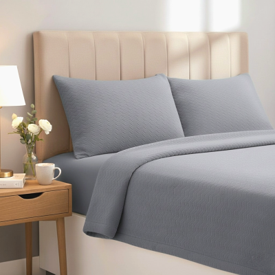 Jogo de cama cinzento matelassê em cama com cabeceira creme e mesa de cabeceira com candeeiro e flores