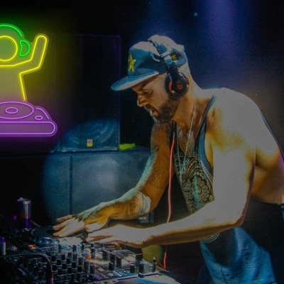 DJ vestido com boné e auscultadores a operar mesa de mistura, luz de neón ao fundo.