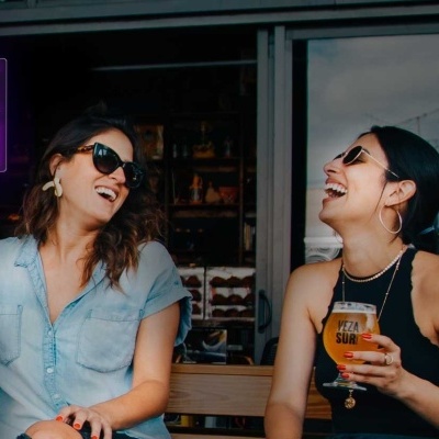 Duas mulheres a rir sentadas num balcão com bebidas ao ar livre, com sinal luminoso rosa a dizer DANCE AGAIN ao fundo.