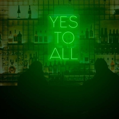 Letreiro de néon verde com texto YES TO ALL num bar com garrafas e pessoas em silhueta.