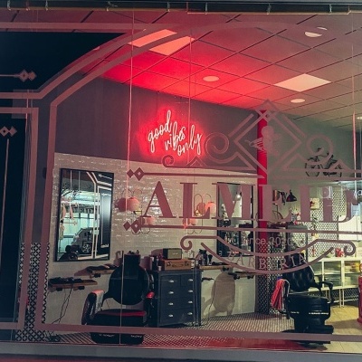 Montra de barbearia com cadeiras pretas, espelhos e luz de néon vermelha com texto 'goodbye beard'