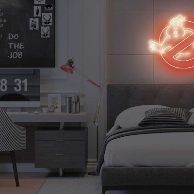 Quarto moderno com cama cinzenta, secretária, cadeira e símbolo neon dos Caça-Fantasmas na parede.