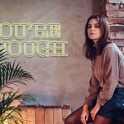 Jovem sentada junto a parede de tijolos e letreiro luminoso com planta verde ao lado