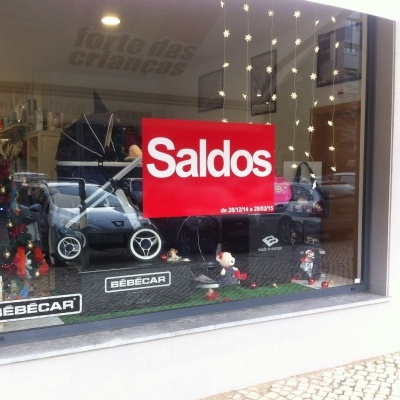 Montra de loja com carrinho de bebé cinzento e placa vermelha com texto 'Saldos'