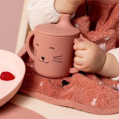 Caneca de silicone cor de rosa com desenho de gato e tampa, segurada por criança com babete rosa