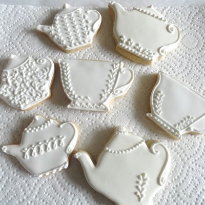 Bolachas decoradas com glacê branco em formas de chávenas e bules sobre toalha de papel branca