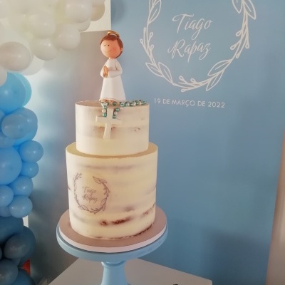 Bolo de comunhão de dois andares com figura de menino no topo e decoração em tons creme e azul