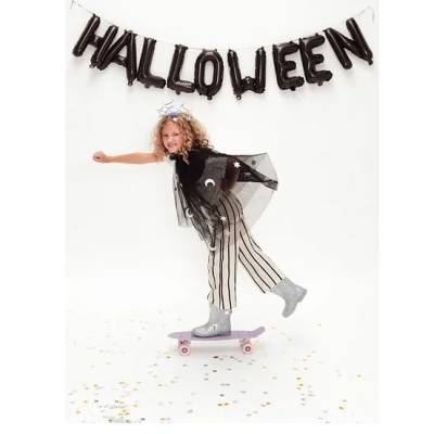 Criança vestida para Halloween com calças às riscas e botas prateadas a andar de skate em fundo branco