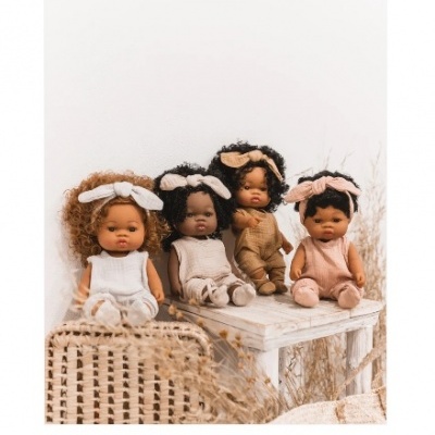 Quatro bonecas de pele castanha clara a escura com roupa de ponto e faixas de cabelo sentadas num banco branco e mala de verga