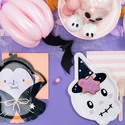 Mesa de festa com pratos decorativos de Halloween, balões cor de rosa e doces sobre fundo lilás.