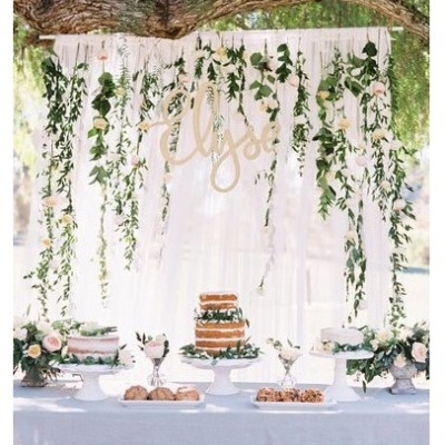 Mesa decorada para festa com bolo, flores e cortina branca com folhas e nome Elyse