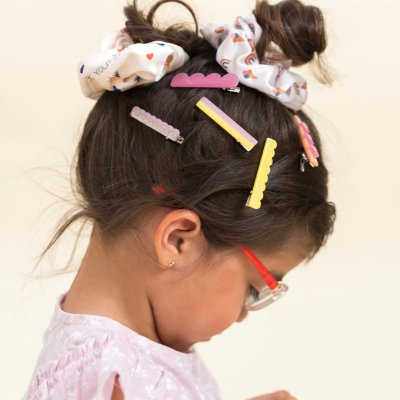 Menina com cabelo preso com ganchos coloridos e scrunchies, óculos vermelhos, blusa rosa clara.
