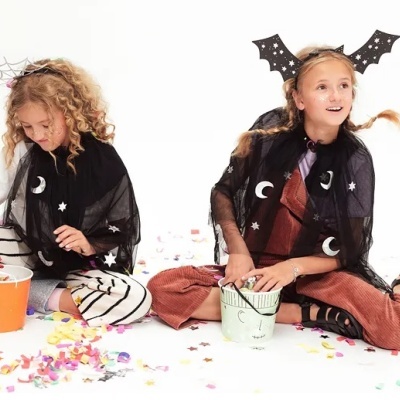 Duas crianças com capas pretas estampadas e calças coloridas sentadas com baldes em chão branco com confetes