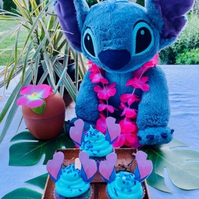 Peluche Stitch azul com colar de flores rosa e cupcakes azuis com corações cor de rosa numa mesa decorada.