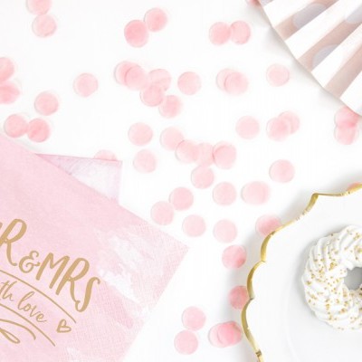 Guardanapos rosa com texto dourado e confetes rosa ao lado de pratos brancos com sobremesa