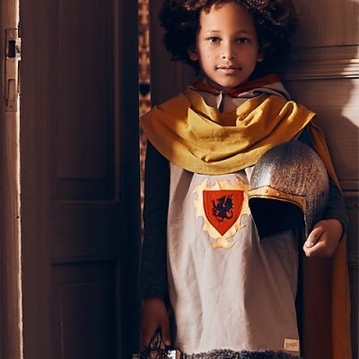 Menino vestido de cavaleiro com capa amarela e capacete metálico junto a porta de madeira.