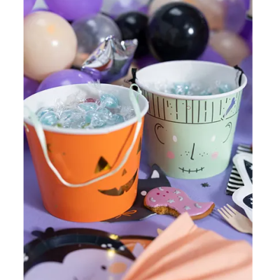 Dois baldes metal laranja e verde com rostos de Halloween numa mesa roxa com balões ao fundo