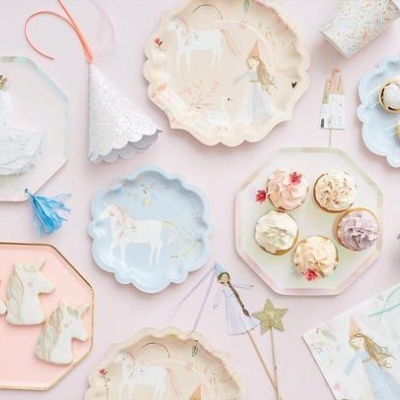 Pratos descartáveis coloridos com desenhos de unicórnios e bailarinas, chapéus de festa brancos e doces sobre fundo rosa.
